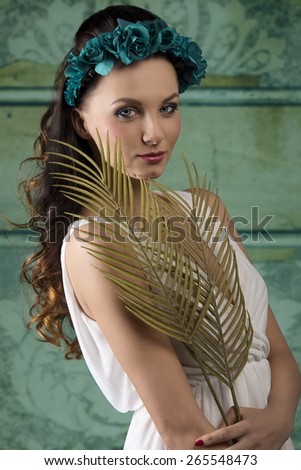 amazing brunette woman with spring style and long wavy hair posing with white dress, some flowers on head and palm leafs in the hands. Romantic expression