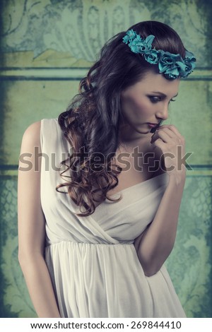 cute brunette female posing in vintage spring portrait with floral crown on the head and sensual white dress, romantic pose
