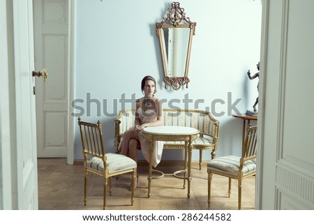 elegant brunette girl in luxury room sitting on vintage sofa with classic hair-style, precious necklace and pink dress. Aristocratic style and antiques furnitures