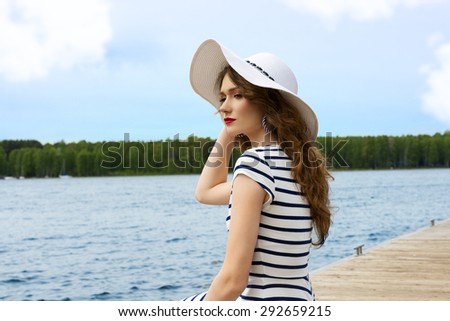 elegant pretty girl , in blue lines dress and summer white hat , outdoor near a lake , with some water on background