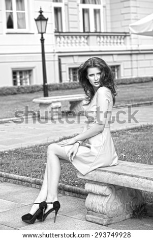 black and white image , very beautiful young woman with elegant pink dress and black heels sitting on old bench in a  park with antique building on background