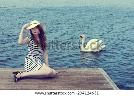 romantic shot of elegant woman in blue lined dress with hat she is sitting on the bridge near the lake with white swan coming
