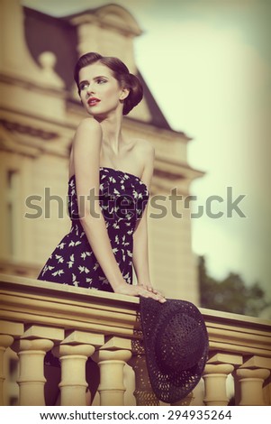 beautiful young brunette girl posing like vintage diva on balcony of ancient palace with elegant hair-style, red lipstick, short dress and hat in the hand .vintage color effect