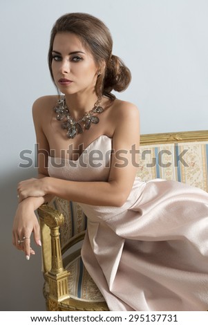 elegant brunette girl with rich style posing with elegant pink dress and precious necklace on old sofa in indoor portrait