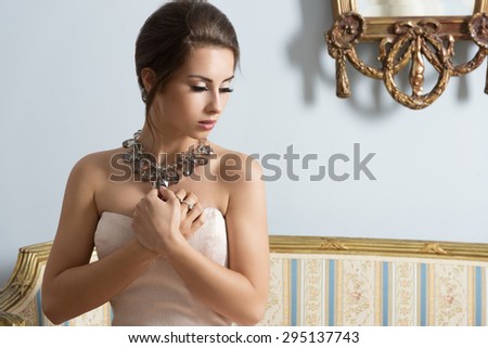 indoor fashion portrait of elegant female with pink dress and big shiny necklace sitting on old sofa in romantic pose