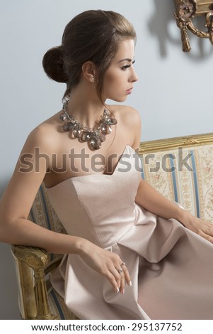 indoor portrait of aristocratic brunette girl sitting on vintage sofa with elegant pink dress, shiny necklace and classic hair-style. Luxury girl in ancient room