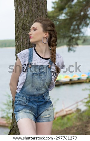 outside portrait of stylish cute woman with braid hair-style, denim overalls and trendy t-shirt posing near tree with relaxed expression and lake water on background.