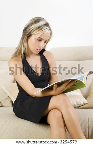 cute and blond girl in black dress reading a magazine and sitting on beige sofa
