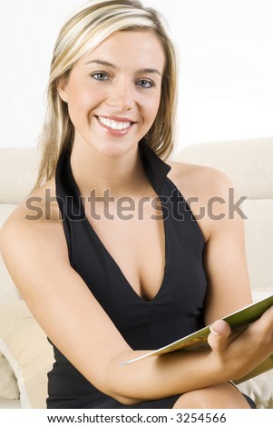 cute and blond girl in black dress reading a magazine and sitting on beige sofa