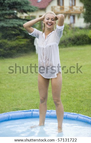 blond girl with wet shirt inside a little swimming pool in a home garden playing with splatter water