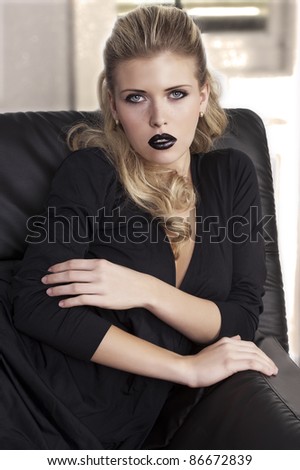 fashion shot of a sophisticated blonde sitting on a black sofa wearing a black dress and black lipstick
