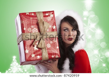 Sexy model in a red christmas dress with short skirt and hood with white fur and a big gift box over light green and christmas trees