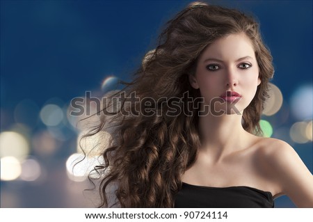 beauty fashion portrait of a very young cute brunette with long curly hair with hairstyle flying in the wind on city light bokeh