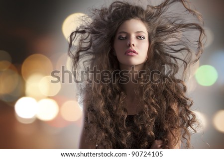 beauty fashion portrait of a very young cute alluring brunette with long curly hair with hairstyle flying in the wind and city lights