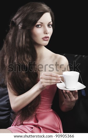 sophisticated young pretty brunette in pink elegant dress sitting on a black sofa and tasting a cup of tea. she looks in to the lens with  three quarters turned faces  and takes a cup of tea