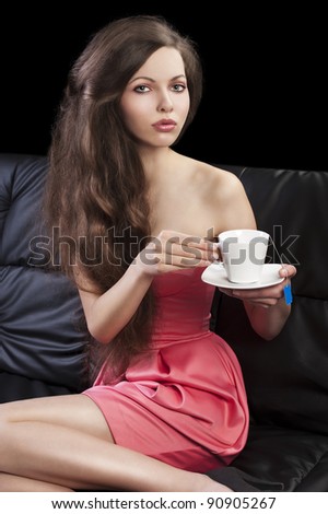 sophisticated young pretty brunette in pink elegant dress sitting on a black sofa and tasting a cup of tea. she looks in to the lens, takes the cup of tea with both hands and her legs are on the sofa.