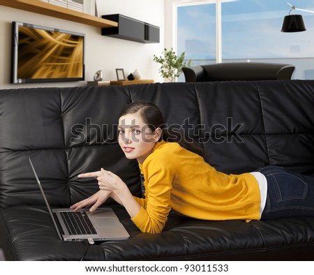 very young girl in yellow shirt laying an a black sofa and working on laptop computer, she\'s liyng on the sofa, looks in to the lens and indicates the laptop\'s display with one finger.