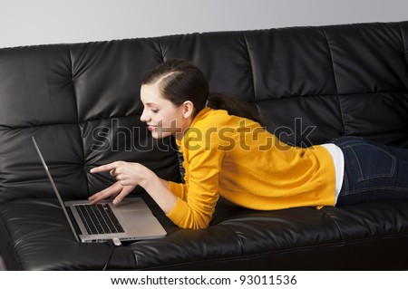 very young girl in yellow shirt laying an a black sofa and working on laptop computer, she\'s liyng on the the sofa, watches the laptop\'s display and indicates that with one finger