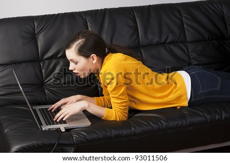 very young girl in yellow shirt laying an a black sofa and working on laptop computer, she\'s liyng on the the sofa, watches the laptop\'s display with expression of surprise