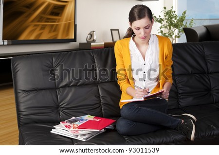 young girl student sitting on a sofa and reading a book making funny face, she\'s on her knees on the sofa, watches the book and take a pen with right hand
