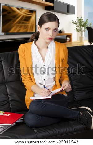young girl student sitting on a sofa and reading a book,she\'s on her knees on the sofa, looks in to the lens and takes a pen with right hand.