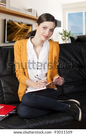 young girl student sitting on a sofa and reading a book with funny face,she\'s on her knees on the sofa, looks in to the lens and smiles, and takes a pen with right hand.