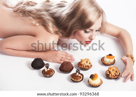 sexy naked woman with long blond hair laying down on white with some pastry near her in act to eat them, she looks pastry and has right hand near one of them