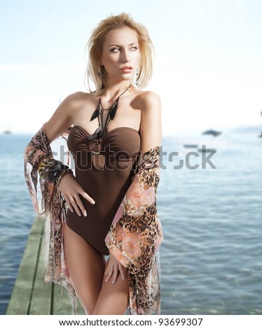 blond sexy beautiful young woman wearing a flower scarf over her body with brown swimsuit with jewellery, she looks at left with sexy expression, her right hand is on the hip and her left hand