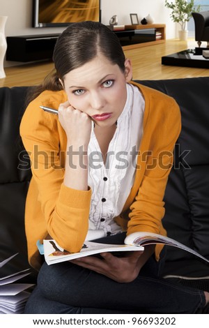 young girl student sitting on a sofa and reading a book, she is on her knees on the sofa, looks on to the lens with serious expression, her face is resting on right fist.