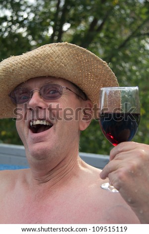 Head & shoulders of middle aged man in cowboy hat with one tooth missing and needing a shave, sitting in hot tub holding a glass of wine with green trees in the background.