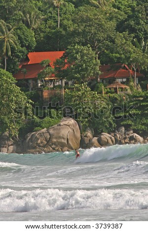 Surfer riding the waves in Phuket in Thailand, with a beautiful backdrop of forest and holiday villa