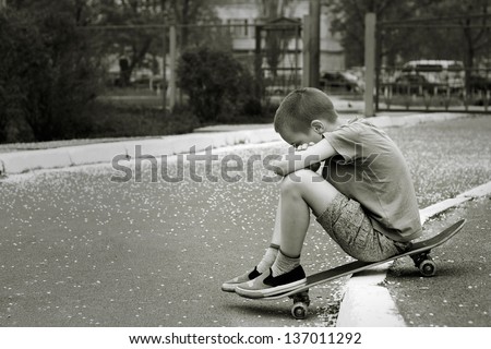 Black and white portrait of sad lonely child who sitting on skateboard in  park