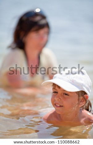 Little girl in baseball hat in the sea and her mother blurred in background