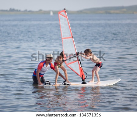 Two little boys try to balance on windsurfing board  with child\'s sail.  Next to them is male instructor