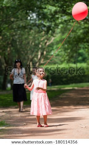 Adorable little girl playing with a red balloon in park. Her family on background