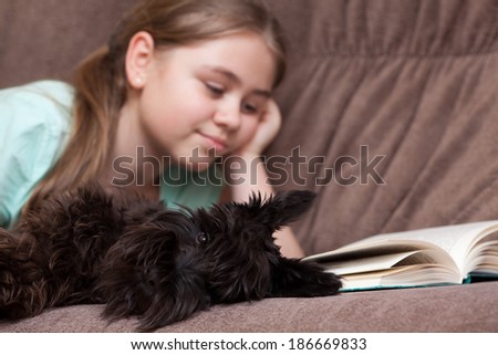 Little girl with dog reading book while lying on the sofa at home. Focus on foreground