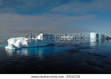 Three Antarctic icebergs in the Southern Ocean on a nearly calm sea covered by ice floes. Underwater parts are visible from the left iceberg.