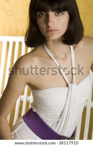 An 18 year old exotic brazilian model, with short dark hair, a young looking face and a skinny body. She is wearing a white dress and purple belt, on a white bench in front of a yellow, textured wall.