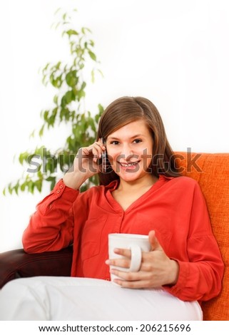 Young woman is sitting on the sofa, having phone conversation and drinking hot drink.