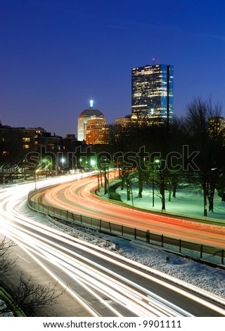 Traffic on Storrow Drive going in and out of downtown Boston at dusk. Car lights lead toward John Hancock Tower.