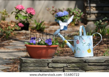 Watering can and flower pot on stone wall, framing path to house entrance. Elegant garden detail.