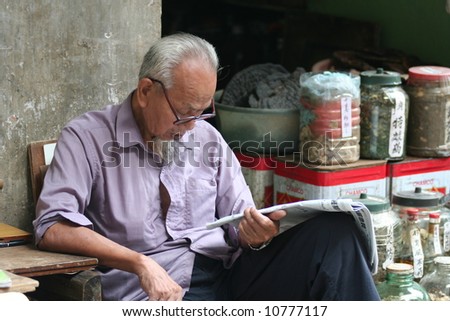 Elderly chinese man reading newspaper in the street in front of his shop