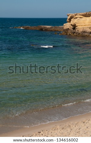 Sand beach with clear water is surrounded with yellow rocks. Sea is tranquil, sky is clear and blue.
