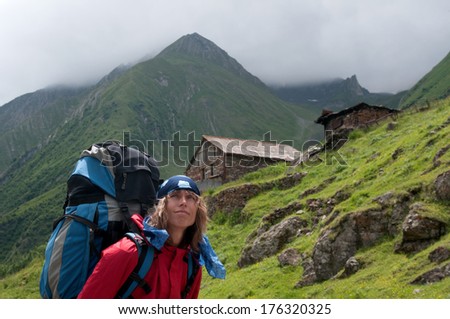 Backpacker (woman) dressed in bright clothes is in the background, mountains covered with clouds are in the background. Little stone houses are on the  mountainside.