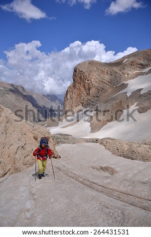 Woman dressed in red windcoat and yellow trousers climbs a mount. Mount slope is covered with snow. Blue sky with clouds and red rocks are in the background.