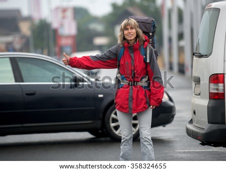 Woman dressed in bright red cloth travels by hitchhiking. Unrecognizable cars are in the background. Woman has a backpack on herself.