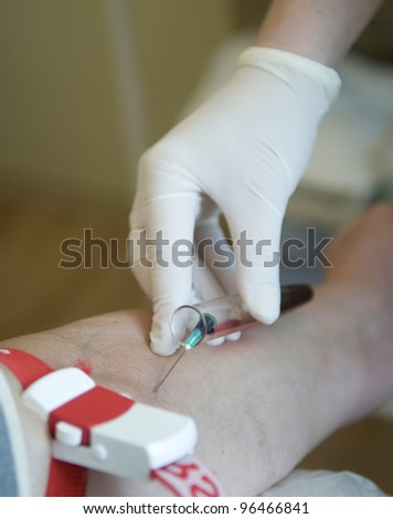 This is a procedure of blood sampling from vein. The gloved hand,  syringe needle, test-glass 	and  patient\'s forearm are in the photograph.