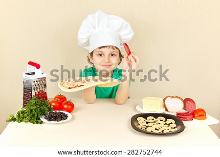 Little boy in chefs hat puts sausage on the pizza crust