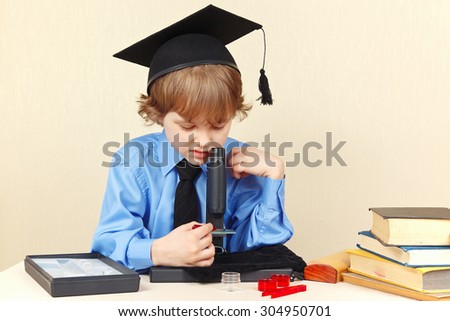 Little serious boy in academic hat looking through a microscope at his desk