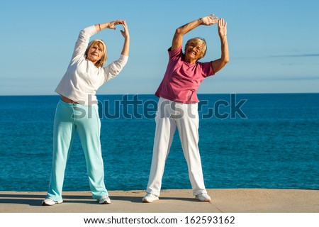 Portrait of senior fitness ladies stretching arms together at seafront.